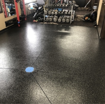 This image shows a gym floor with speckled rubber matting. There are dumbbells lined against the wall, and a person is partially visible lying down.
