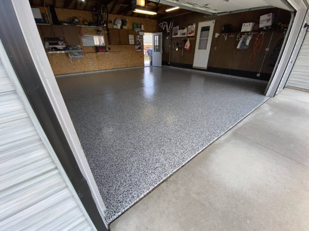A tidy garage interior with an epoxy-coated floor, a workbench, shelving, and various tools and equipment neatly arranged along the walls. Garage door is open.