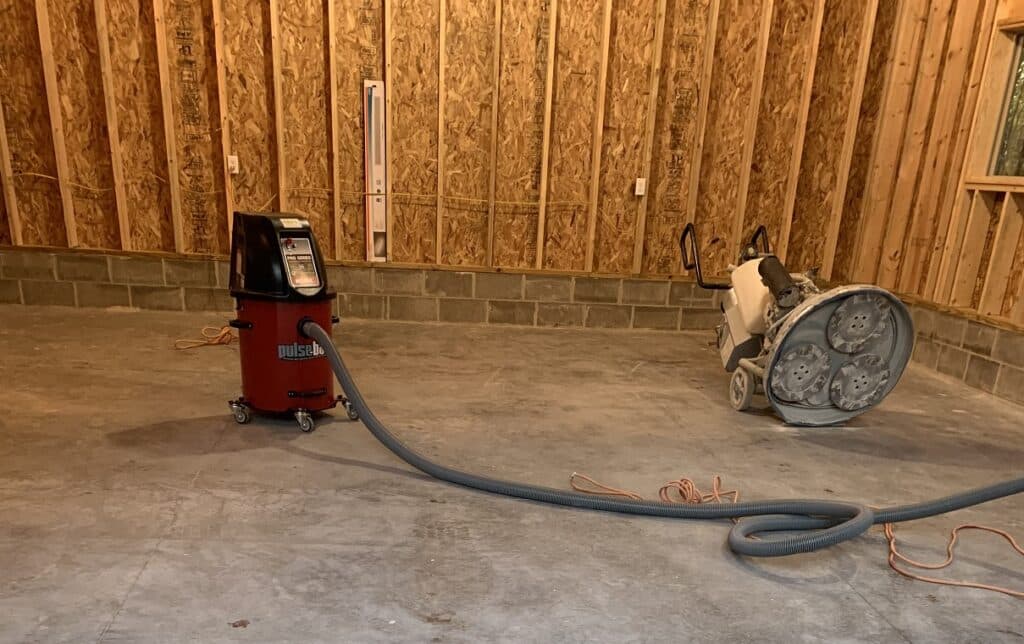 The image shows a construction interior with wooden walls. There's a red industrial vacuum cleaner and a concrete floor grinder on a concrete floor.