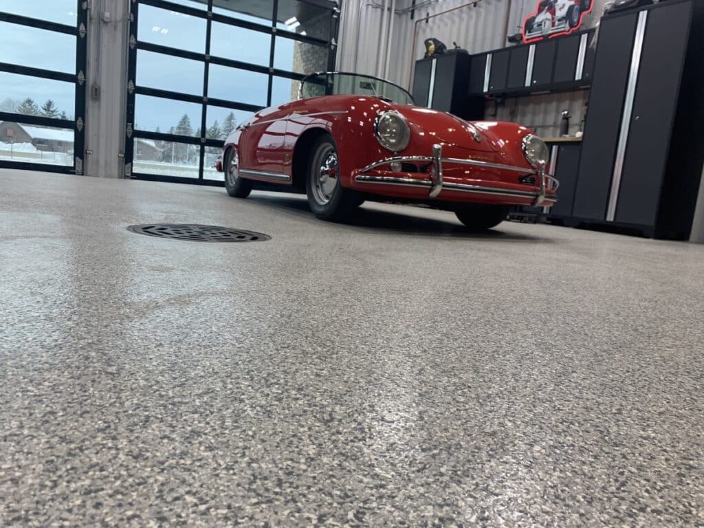 A classic red convertible car is parked inside a garage with large windows, on a speckled floor, near a workspace with cabinets and a wall art piece.