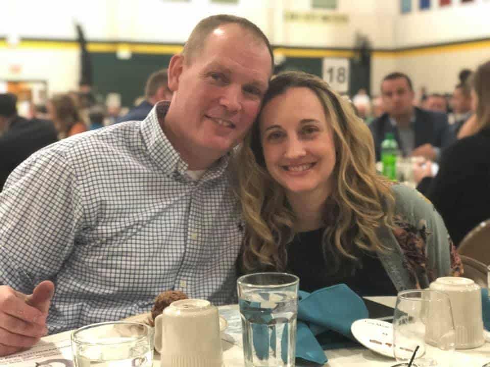 Two people smiling at a table with dinnerware, at a social event with blurry guests in the background. They appear cheerful and close together.
