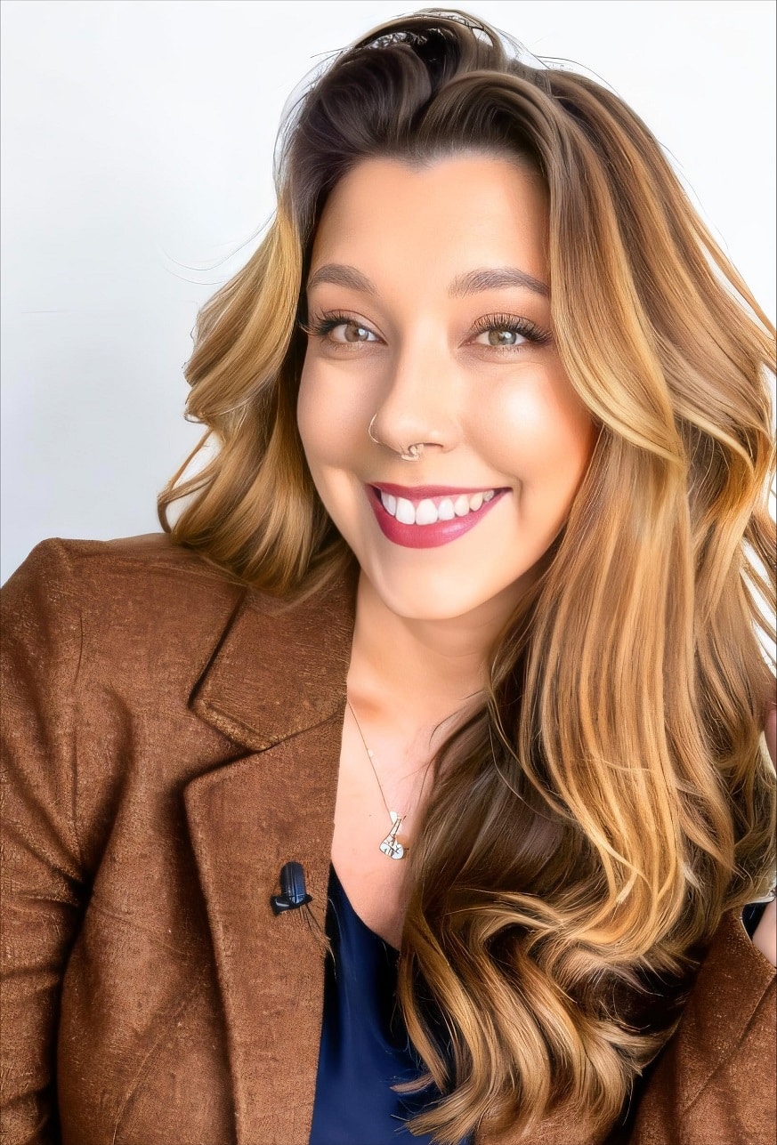 A smiling person with a nose ring, long wavy hair, wearing a brown blazer over a navy top, and a necklace, against a white background.