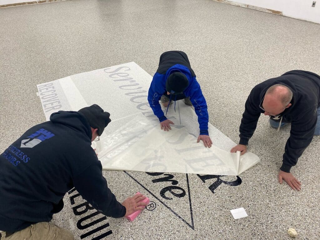 Three people are working together to apply a large adhesive decal to a textured floor, concentrating carefully on their task.