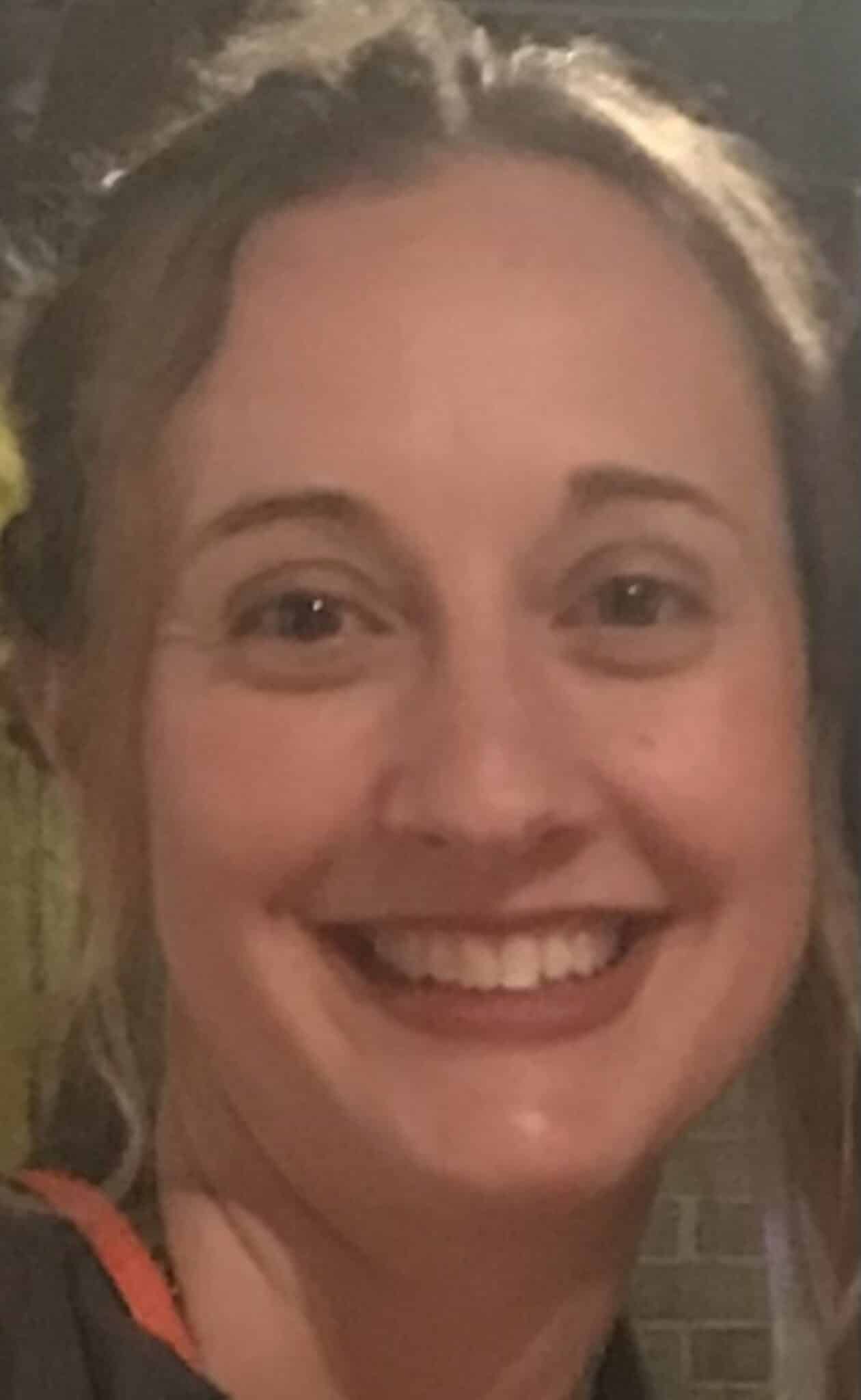 Close-up of a smiling person with light skin, cheerful expression, visible teeth, sparkling eyes, and a blurred background.