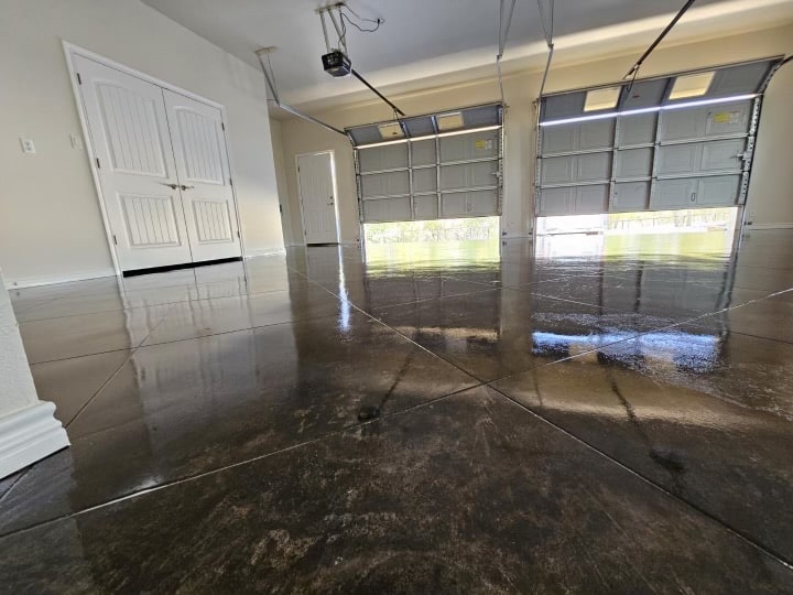 An empty garage with a shiny, polished concrete floor reflecting the surrounding interior, including dual white doors and an open overhead door revealing daylight.
