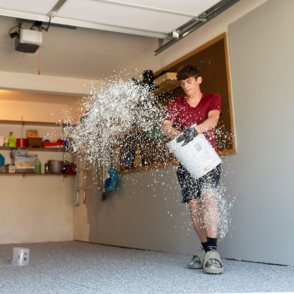 A person in a garage spills a multitude of small white pellets mid-air from a large bucket, creating a dynamic and messy scene.
