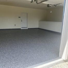 This is an image of an empty garage with a speckled floor coating, walls painted in a light color, and a white door at the back.