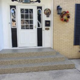 This is the front entrance of a house with a white door, yellow brick facade, decorative wreath, window shutters, and a set of steps.