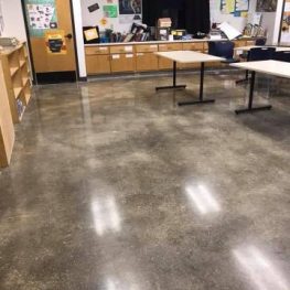 The image shows an indoor setting with polished concrete floors, office furniture including tables and desks, and informative boards on the walls.