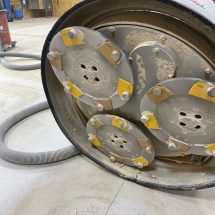 Grinder with Diamonds This image shows a close-up of a heavy-duty industrial grinding machine with diamond-segmented wheels, against a plywood wall and concrete floor, with a flexible hose nearby.