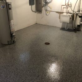 This image shows a clean utility room with a water heater, white walls, and speckled grey flooring with a drain. Various pipes and a laundry basin are visible.