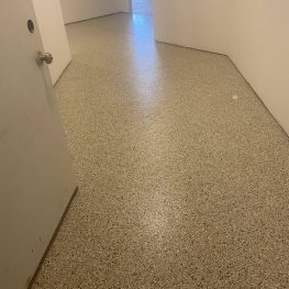 This image shows a corridor with a speckled floor, white walls, a door on the left, and a small trash bin visible at the far end.