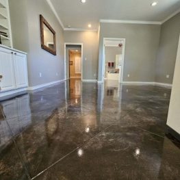An empty room with polished brown tile flooring, white walls, and several doorways leading to other rooms. A staircase and a framed picture are visible. Light streams through a window.