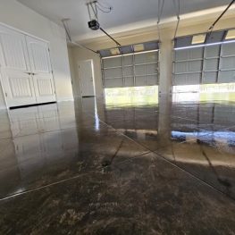 An empty garage with a shiny, polished concrete floor reflecting the surrounding interior, including dual white doors and an open overhead door revealing daylight.